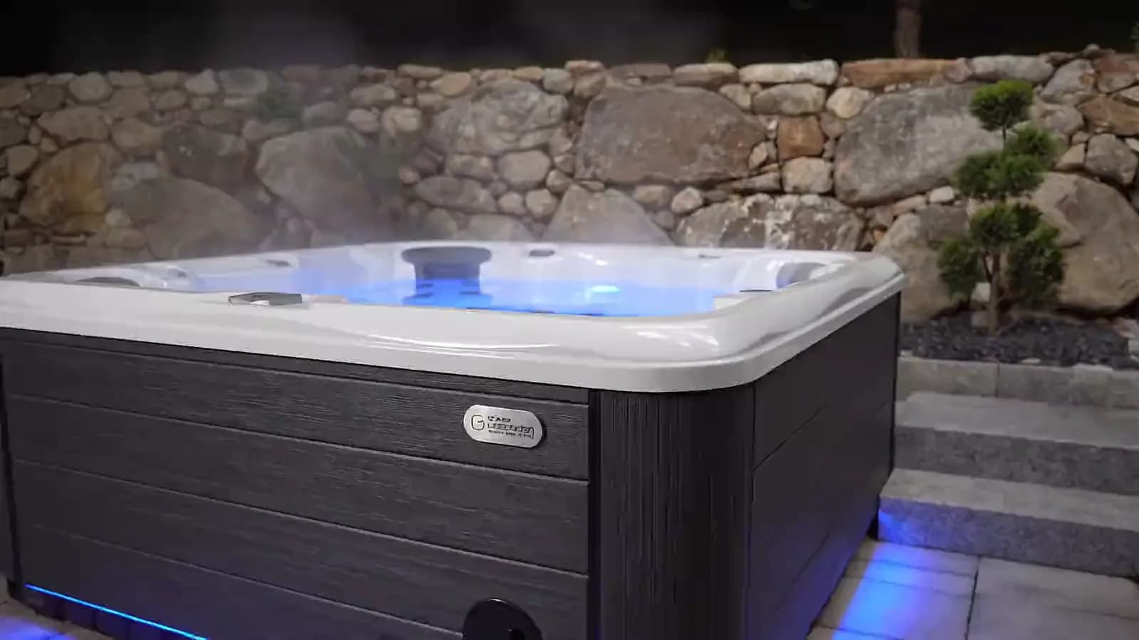 Spa éclairé de nuit sur une terrasse en pierre, vapeur au-dessus de l'eau, muret de pierres sèches et jardin éclairé en arrière-plan