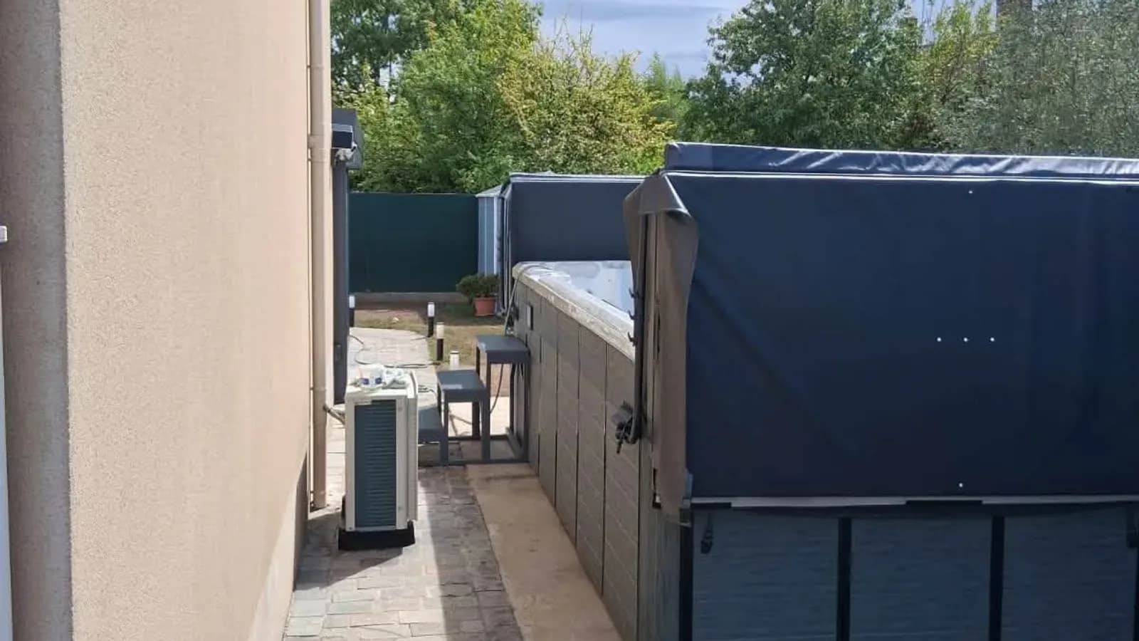 Spa de nage Oxford installé le long d'une maison à Forges-les-Bains, couverture fermée, avec sa pompe à chaleur et sa terrasse en pierre